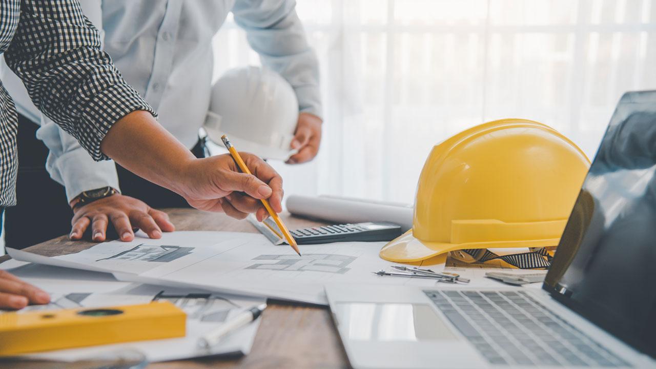 An open laptop, yellow hard hat, and design documents on a table with people gathered around reviewing the documents